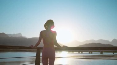Ahşap iskelede yürüyen ve dağlarla ve deniz manzaralı gün batımını izleyen kadın silueti. Kadın akşam antrenmanından sonra dinleniyor.