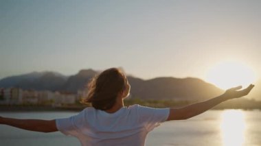 Happy carefree woman turning around to camera with hand, enjoying sunset time with orange sky. Sundown landscape with mountains and sea view