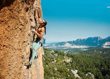 Güçlü kaslı vücut adamı kaya tırmanışçısı dağlarda ip ve mavi gökyüzü arka planıyla dikey kayaya tırmanıyor. Açık hava sporları ve eğlence