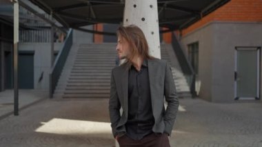 Sakallı ve uzun saçlı yakışıklı adam ofis arka planında dışarıda duruyor. Gri ceket ve kot pantolon giyiyor. Yana bakıyor. Adam rüya gibi görünüyor.