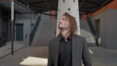 Sakallı ve uzun saçlı yakışıklı adam ofis arka planında dışarıda duruyor. Gri ceket ve kot pantolon giyiyor. Yana bakıyor. Adam rüya gibi görünüyor.