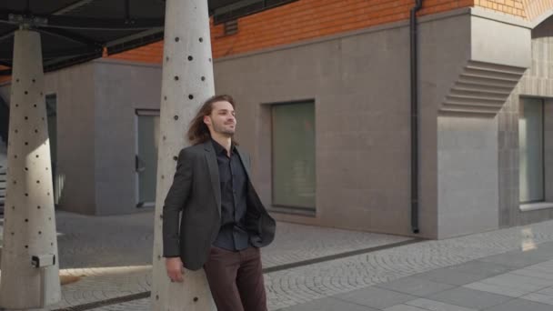 一个留着胡子和长发的英俊男子正站在办公大楼背景的室外 他穿着灰色夹克 牛仔裤 他向侧面看去 看起来很有梦想的男人 — 图库视频影像