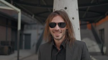 Sakallı ve uzun saçlı yakışıklı adam ofis arka planında dışarıda duruyor. Gri ceket ve kot pantolon giyiyor. Yana bakıyor. Adam rüya gibi görünüyor.