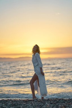 Sahilde günbatımının tadını çıkaran, şık elbiseli çekici genç bir bayan. Arka plan turuncu gün batımı Denizde
