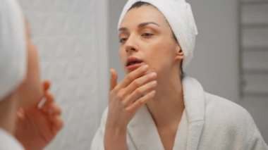 Beyaz banyo havlusuna sarılı, parlak banyoda duran ve parmaklarıyla yüz masajı yapan çarpıcı genç bayan. Mutlu kadın cildinin halinden memnun..