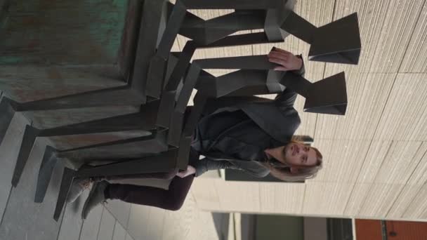 スーツ姿の垂直ビデオ孤独なビジネスマンがオフィスビルの近くに座って巨大な金属人間の彫刻 金融街の屋外で一人でビジネスをしていても — ストック動画