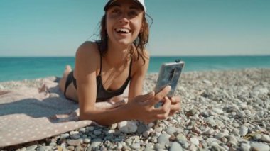 Rahat kadın turist deniz kıyısında güneşlenir, cep telefonunun kilidini açar ve fotoğraf ya da video çeker. Mavi Göl beldesi, Oludeniz, Türkiye.