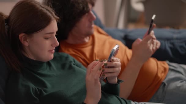 仕事の後若い夫婦は自宅のソファに横になり両方ともスマートフォンを見ています 人々は一緒に時間を過ごすだけでなく自分の携帯電話やSnsやメッセンジャーを — ストック動画