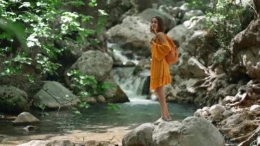 Turuncu elbiseli genç kadın dağ nehri boyunca kaya parçalarıyla yürüyor ve ormanda yürüyüş yaparken etrafına bakıyor. Vahşi ormanda yürüyüş sıcak yaz günü, açık hava aktivitesi ve vahşi doğada seyahat.