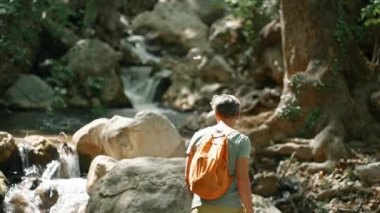 Ağır çekim yürüyüşçüsü dağ nehri boyunca kaya parçalarıyla yürüyor ve ormanda yürüyüş yaparken etrafına bakıyor. Sıcak yaz gününde vahşi ormanda yürüyüş, açık hava aktivitesi ve seyahat konsepti. İnsanlar ve vahşi doğa