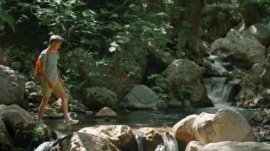 Ağır çekim yürüyüşçüsü dağ nehri boyunca kaya parçalarıyla yürüyor ve ormanda yürüyüş yaparken etrafına bakıyor. Sıcak yaz gününde vahşi ormanda yürüyüş, açık hava aktivitesi ve seyahat konsepti. İnsanlar ve vahşi doğa