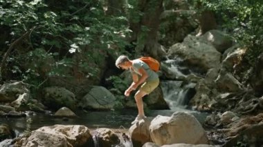 Ağır çekim yürüyüşçüsü nehirde kayalarla yürüyor ve ormanda yürüyüş yaparken dereden taze soğuk su içiyor. Sıcak yaz gününde vahşi ormanda yürüyüş, açık hava aktivitesi ve seyahat konsepti.