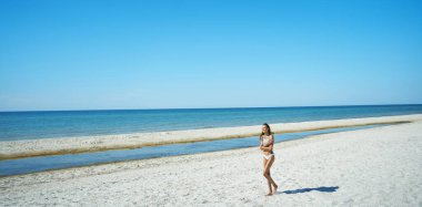 Cennet gibi bir sahil kaçamağının güzel panoramik görüntüsü. Güneş yanığı kadın, deniz manzaralı sakin kumlu sahilde yürüyor.