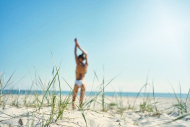 Göz kamaştırıcı güneşli bir plajın huzur görüntüsü. Arka planda, beyaz kumlu deniz kıyısında kollarını mavi gökyüzüne doğru uzatan kadının odaklanmamış görüntüsü var.
