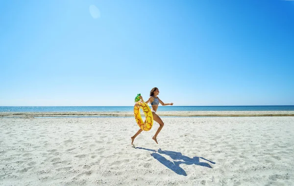美しいパノラマ画像の楽園の風景 無限の砂浜に沿って実行されているブロンズ喜びの若い女性 パイナップルフロートリングを運ぶ 紺碧の水に飛び込む準備ができて — ストック写真