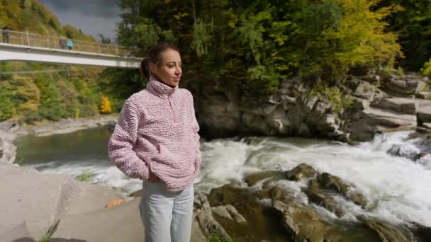 轻松自在的女游客站在靠近山河的岩石上 瀑布重重 秋山远足时 女徒步旅行者欣赏大自然的美丽 户外运动休闲 享受大自然 — 图库视频影像