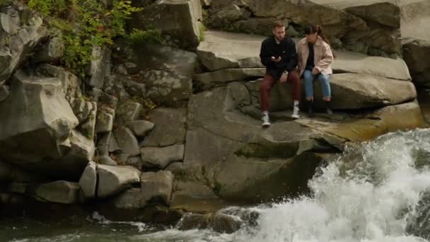 快乐的情侣游客们坐在靠近山河的岩石上 瀑布重重 秋天登山时 可爱的男女徒步旅行者欣赏大自然的美丽 户外运动休闲 享受大自然 — 图库视频影像