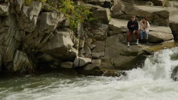 快乐的情侣游客们坐在靠近山河的岩石上 瀑布重重 秋天登山时 可爱的男女徒步旅行者欣赏大自然的美丽 户外运动休闲 享受大自然 — 图库视频影像