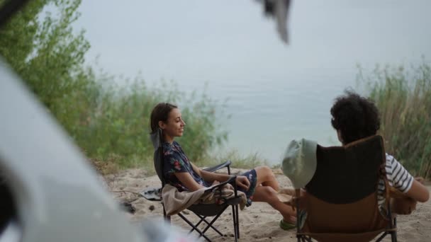 两个朋友坐在营地的椅子上 靠近湖边的野营车 在沙滩上聊天 夫妻二人在湖边野营时外出野餐 — 图库视频影像