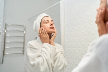 Beyaz banyo havlusuna sarılı, parlak banyoda duran ve parmaklarıyla yüz masajı yapan çarpıcı genç bayan. Mutlu kadın cildinin halinden memnun..