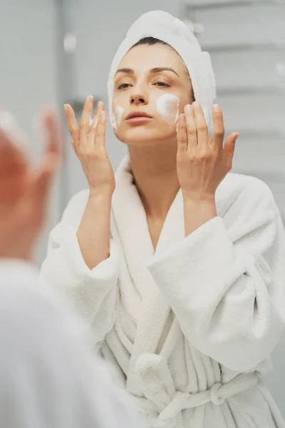 女人用保湿泡沫洗脸 晨光护肤 成年女性头戴毛巾 在浴室照镜子 化妆的准备工作 — 图库照片