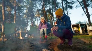 Sıcak yürüyüş yapan iki kadın akşam vakti Güz Ormanı 'nda Şenlik Ateşi' nin yanında otururken dumandan yanıyor. Doğadaki şömine - geniş açılı çekim, yavaş çekim. piknik ve açık havada kamp.
