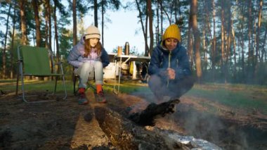 Sıcak yürüyüş yapan iki kadın akşam vakti Güz Ormanı 'nda Şenlik Ateşi' nin yanında otururken dumandan yanıyor. Doğadaki şömine - geniş açılı çekim, yavaş çekim. piknik ve açık havada kamp.