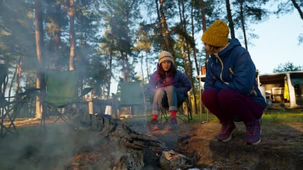 两名穿着保暖衣服的女徒步旅行者坐在篝火边 黄昏时分在秋天的森林里被烟熏着 火焰在自然界广角镜头 慢动作 野外野餐和野营 — 图库视频影像