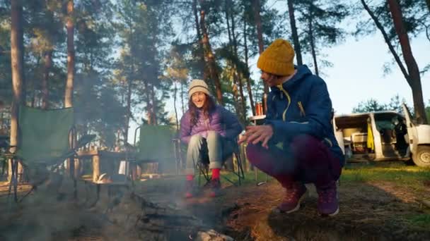 两名穿着保暖衣服的女徒步旅行者坐在篝火边 黄昏时分在秋天的森林里被烟熏着 火焰在自然界广角镜头 慢动作 野外野餐和野营 — 图库视频影像