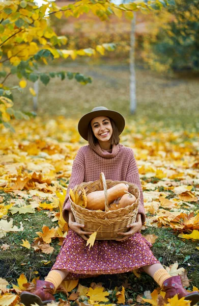 Young happy country woman in autumnal stylish wear holding wicker basket filled with freshly ripe pumpkins. Nature beauty of fall season and autumnal harvest