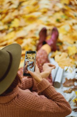 Güzel bir sonbahar parkında dinlenirken şapkalı bir kadının kameralı telefonuyla fotoğraf çekişi. Akıllı telefondaki resme odaklan