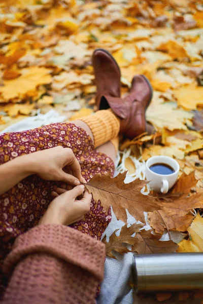 Trendy autumn fashion and atmospheric mood. Legs in vintage boots and yellow socks of women in red dress lying surrounded fall leaves during coffee picnic outdoors