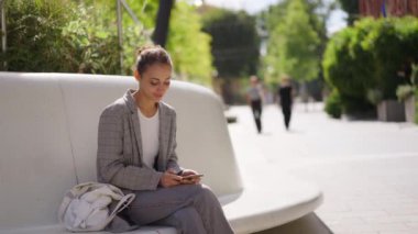 Şehir meydanında taştan bir bankta oturan zarif melez genç bir kadın. Birini bekliyor ve telefonunu kullanıyor. kız cep telefonu ekranına bakıyor, arıyor, yazıyor.