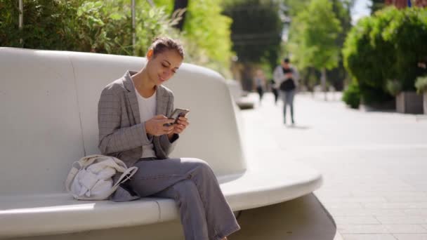 优雅的年轻的混血女子坐在城市广场的石凳上等着别人 并使用她的电话 穿着灰色夹克和牛仔裤的女孩看上去像手机屏幕 — 图库视频影像