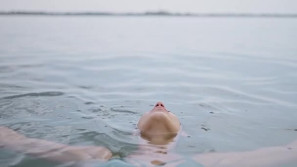 一位年轻的成年女子笑着放松地躺在湖面上 眼睛心满意足地闭上眼睛 享受着泡在水里的乐趣 站在水面上看着相机的女人 — 图库视频影像