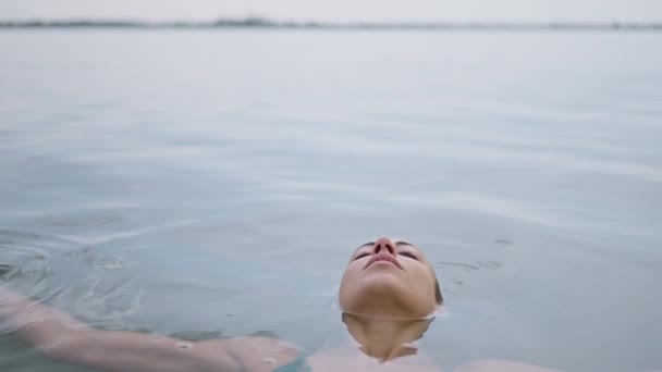 一位年轻的成年女子放松地躺在湖面上 心满意足地闭上眼睛 享受着泡在水里的乐趣 站在水面上看着相机的女人 — 图库视频影像