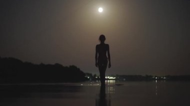 Sıska bir kadının gölde gece vakti ay ışığında sığ suda yürümesi. Yaz gecesi karanlıkta yüzen şehvetli kız