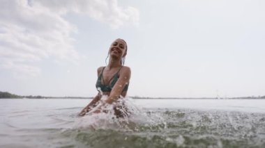 Yeşil mayo giymiş, göl suyunda eğlenen ve elleriyle su sıçratan güzel, neşeli, genç seksi kız. Plajda tropik arka planda bikinili güzel bir kadın vücudu.