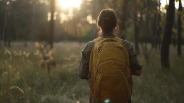 Kamera, güzel ormanda yürüyen ve güzel doğayı keşfeden genç bir kadını takip ediyor. Sarı sırt çantalı ve tişörtlü. Kamerada güneş parlıyor, mercek parlıyor..