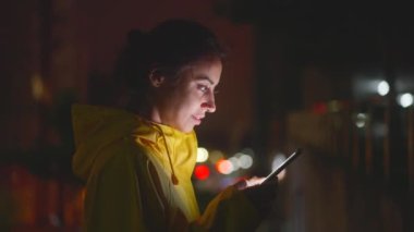 Sarı yağmurluklu gülümseyen kadın iletişim kurmak için akıllı telefon kullanıyor. Gece caddelerinde yürüyen, telefonda yazan güzel bir kadın. Sosyal Ağ ve Çevrimiçi İletişim Konsepti. Yavaş Hareket Görüntüsü