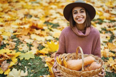 Şapkalı neşeli kız kameraya bakıyor ve elinde olgun balkabaklarıyla hasır sepet tutuyor. Sonbahar mevsiminin ve sonbahar hasadının doğa güzelliği