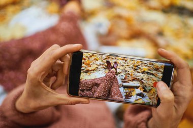 Akıllı telefonu tutan ellerini kapat ve turuncu yapraklarla çevrili piknik sırasında bacaklarının fotoğrafını çek.
