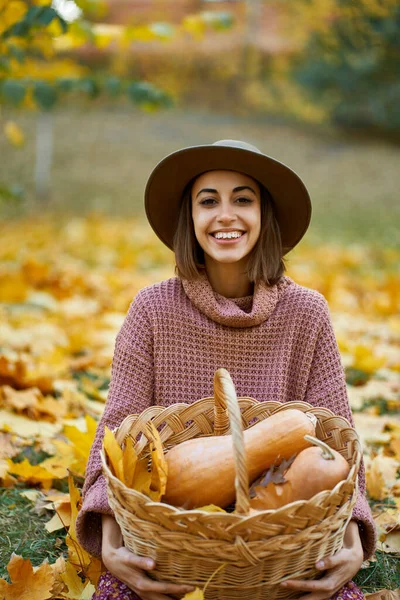 Portrait of happy country woman in autumnal stylish wear holding wicker basket filled with freshly ripe pumpkins. Nature beauty of fall season and autumnal harvest