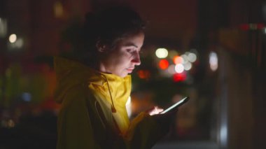 Sarı yağmurluklu kadın şehrin dışında akıllı telefon kullanıyor. Gece sokakta yürüyen, telefonda yazan ve ekrana bakan bir kadın. Sosyal Ağ ve Çevrimiçi İletişim Kavramı.