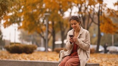 Sonbaharda parkta şubede otururken akıllı telefon kullanan mutlu, güzel, genç bir kadın. Çekici, sevimli, neşeli kadın dışarıda internet teknolojilerini kullanıyor. Akıllı telefon kavramı.