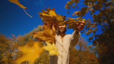 Merhaba sonbahar. Sonbaharda şehir parkında açık havada neşe içinde gülümseyen, açık ceketli modern bir kadın. Kadın kuru akçaağaç yaprağı kusar ve güler.