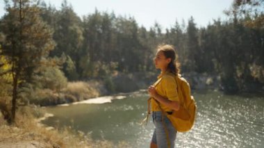 Güneşli bir sonbahar sabahı, sıcak bir günde, çam ormanında dağ gölü etrafında yürüyüş yapan kadın yürüyüşçüyü takip ediyoruz. Sarı sırt çantalı kadın gezgin ormanda yürüyüş yapıyor, maceracı açık hava yaşam tarzı.