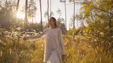 Güzel, mutlu, beyaz elbiseli bir kadın sonbahar ormanında uzun, kuru çimenlerin altın dikenleri arasında neşeyle yürüyor. Romantik kız büyülü sonbahar ormanlarında dinleniyor.