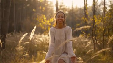 Güzel, mutlu, beyaz elbiseli bir kadın sonbahar ormanında uzun, kuru otların altın dikenleri arasında neşeyle yürüyor. Romantik kız büyülü sonbahar ormanlarında dinleniyor.