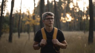 Yavaş çekim yetişkin gri saçlı, gözlüklü, sarı sırt çantalı adam sonbahar ormanında yol kenarında yürüyor. Sırt çantalı erkek yürüyüşçü yüksek otlarla ağaçların arasından yukarı çıkıyor. Seçici odaklanma. Ukrayna 'da seyahat